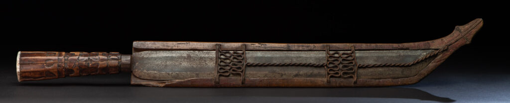 Paiwan tribe hunting knife with a sheath carved with a standing figure and human faces on the side. The hilt is made of carved wood.