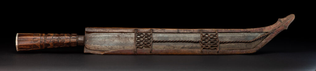 Paiwan tribe hunting knife with a sheath carved with a standing figure and human faces on the side. The hilt is made of carved wood.