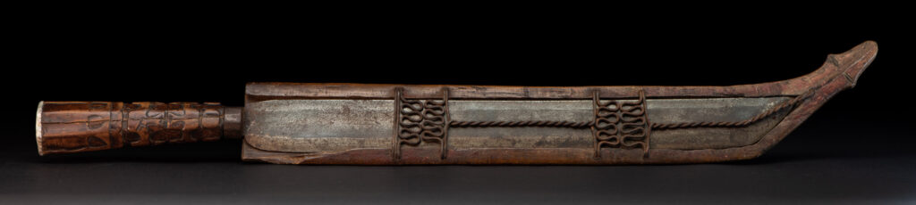 Paiwan tribe hunting knife with a sheath carved with a standing figure and human faces on the side. The hilt is made of carved wood.