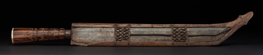 Paiwan tribe hunting knife with a sheath carved with a standing figure and human faces on the side. The hilt is made of carved wood.
