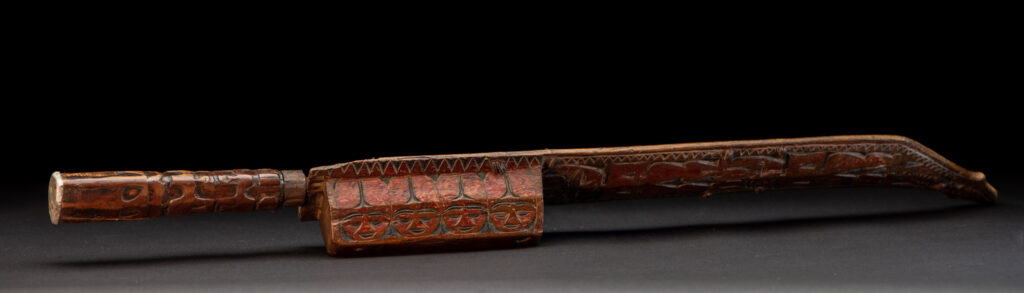 Paiwan tribe sword with a sheath carved with a standing figure and human faces on the side, peint in red. The hilt is made of carved wood.