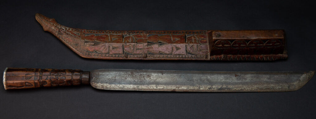 Paiwan tribe sword with a sheath carved with a standing figure and human faces on the side, peint in red. The hilt is made of carved wood.