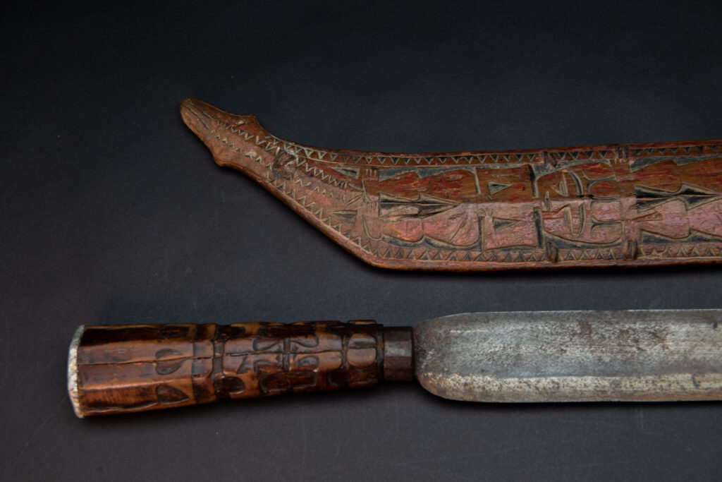 Paiwan tribe sword with a sheath carved with a standing figure and human faces on the side, peint in red. The hilt is made of carved wood.