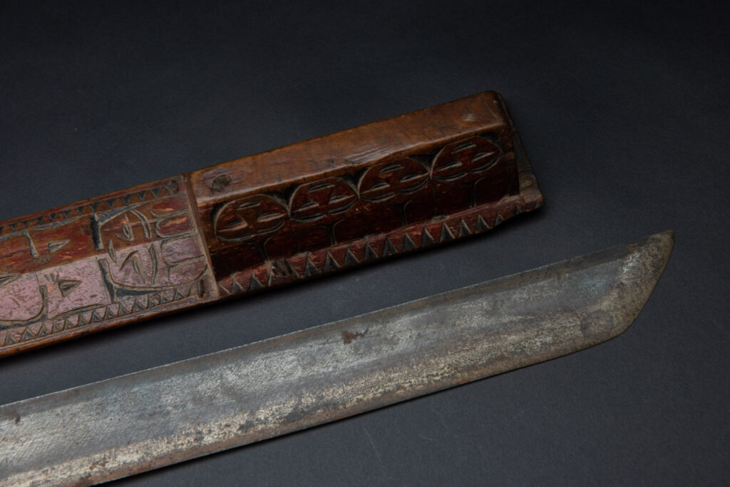 Paiwan tribe sword with a sheath carved with a standing figure and human faces on the side, peint in red. The hilt is made of carved wood.