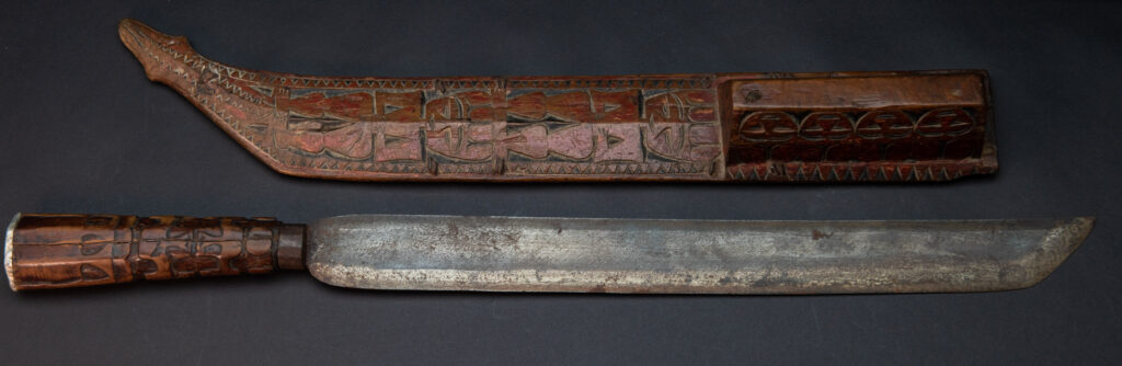 Paiwan tribe sword with a sheath carved with a standing figure and human faces on the side, peint in red. The hilt is made of carved wood.
