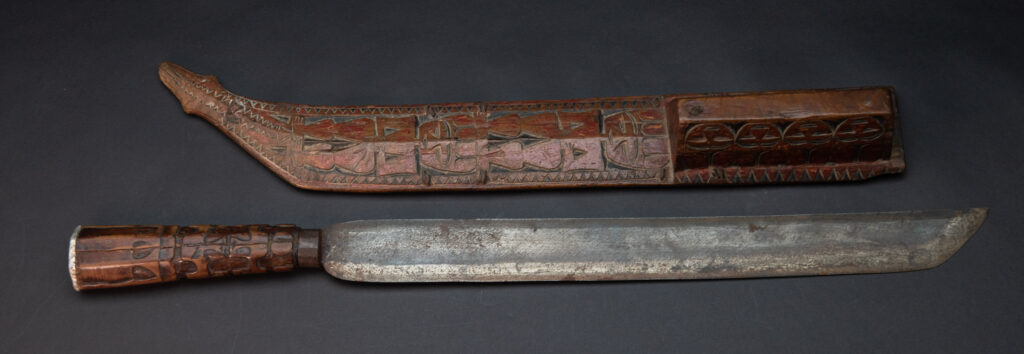 Paiwan tribe sword with a sheath carved with a standing figure and human faces on the side, peint in red. The hilt is made of carved wood.