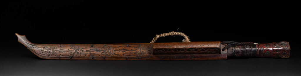 Very fine ceremonial sword from the Paiwan tribe, featuring a sheath carved with full-standing ancestral figures and a handle tinted with red pigment. The sides are carved with human faces.