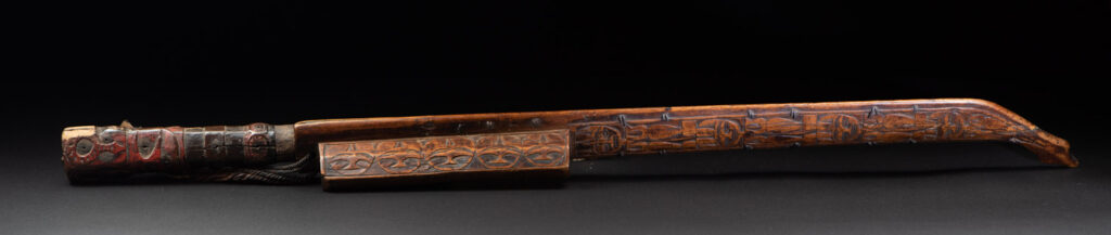 Very fine ceremonial sword from the Paiwan tribe, featuring a sheath carved with full-standing ancestral figures and a handle tinted with red pigment. The sides are carved with human faces.