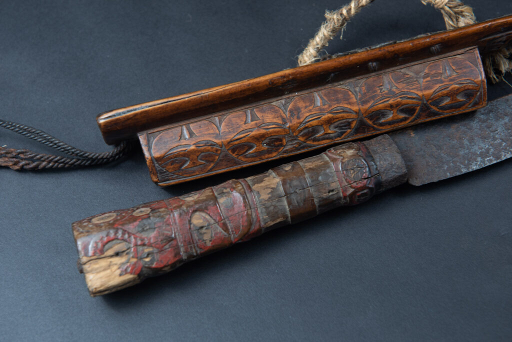 Very fine ceremonial sword from the Paiwan tribe, featuring a sheath carved with full-standing ancestral figures and a handle tinted with red pigment. The sides are carved with human faces.