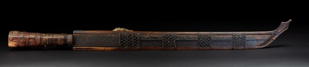 Very fine ceremonial sword from the Paiwan tribe, featuring a sheath carved with full-standing ancestral figures and a handle tinted with red pigment. The sides are carved with human faces.