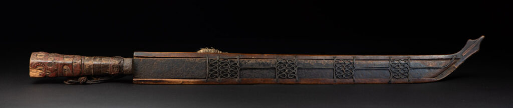 Very fine ceremonial sword from the Paiwan tribe, featuring a sheath carved with full-standing ancestral figures and a handle tinted with red pigment. The sides are carved with human faces.