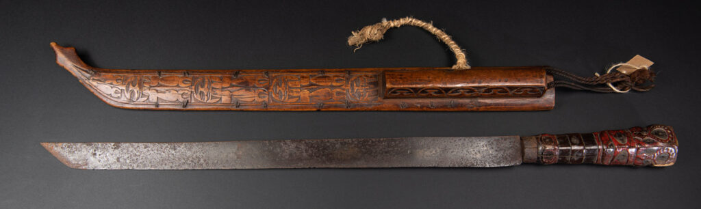Very fine ceremonial sword from the Paiwan tribe, featuring a sheath carved with full-standing ancestral figures and a handle tinted with red pigment. The sides are carved with human faces.