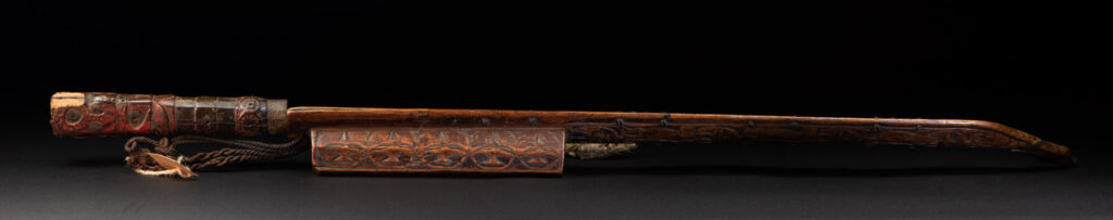 Very fine ceremonial sword from the Paiwan tribe, featuring a sheath carved with full-standing ancestral figures and a handle tinted with red pigment. The sides are carved with human faces.