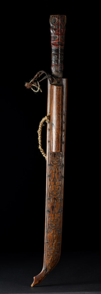 Very fine ceremonial sword from the Paiwan tribe, featuring a sheath carved with full-standing ancestral figures and a handle tinted with red pigment. The sides are carved with human faces.