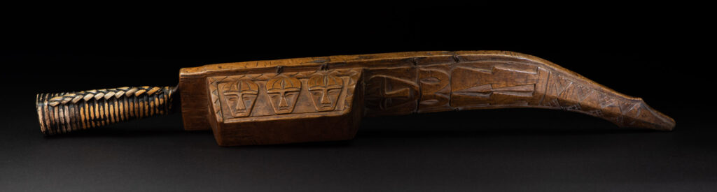 Short sword from the Paiwan tribe, with a sheath carved with a standing figure. The side of the sheath is adorned with three sculpted human heads. The sword is made in one piece with the handle, which is wrapped in rattan.