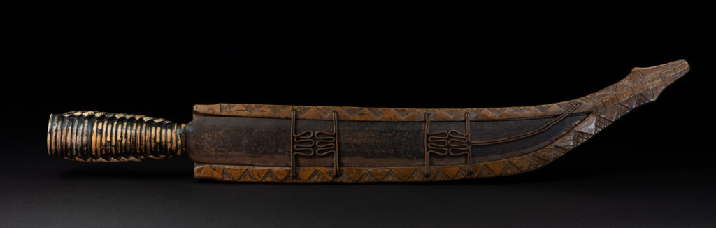 Short sword from the Paiwan tribe, with a sheath carved with a standing figure. The side of the sheath is adorned with three sculpted human heads. The sword is made in one piece with the handle, which is wrapped in rattan.