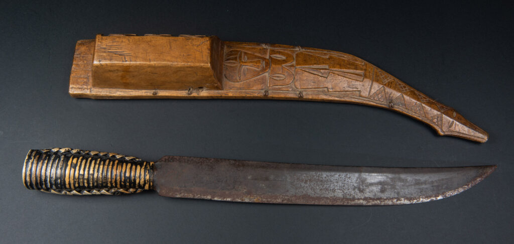 Short sword from the Paiwan tribe, with a sheath carved with a standing figure. The side of the sheath is adorned with three sculpted human heads. The sword is made in one piece with the handle, which is wrapped in rattan.