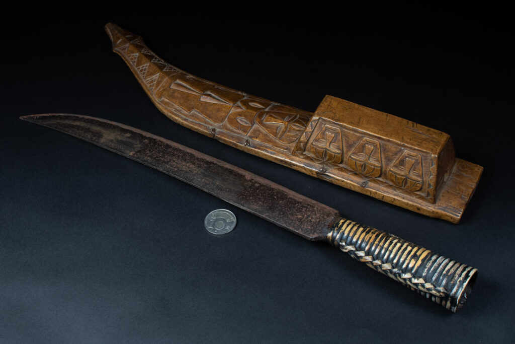 Short sword from the Paiwan tribe, with a sheath carved with a standing figure. The side of the sheath is adorned with three sculpted human heads. The sword is made in one piece with the handle, which is wrapped in rattan.