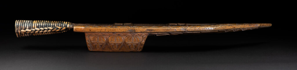 Short sword from the Paiwan tribe, with a sheath carved with a standing figure. The side of the sheath is adorned with three sculpted human heads. The sword is made in one piece with the handle, which is wrapped in rattan.