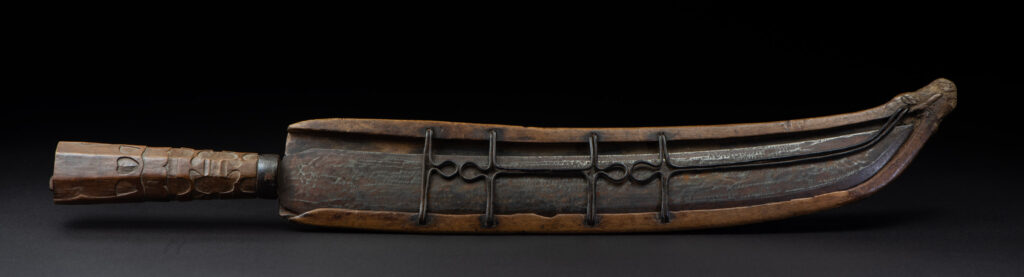 Paiwan tribe hunting knife with a sheath carved with a standing figure and human faces on the side. The hilt is made of carved wood.