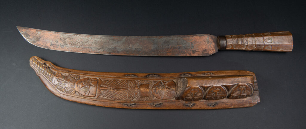 Paiwan tribe hunting knife with a sheath carved with a standing figure and human faces on the side. The hilt is made of carved wood.