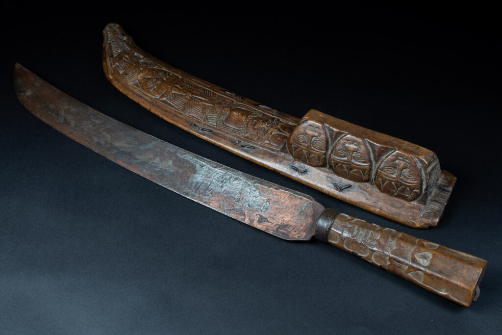 Paiwan tribe hunting knife with a sheath carved with a standing figure and human faces on the side. The hilt is made of carved wood.