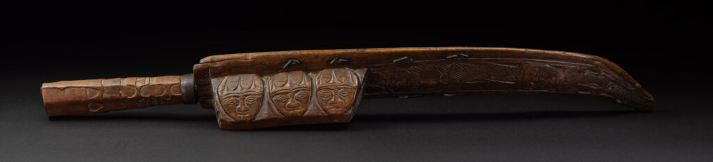 Paiwan tribe hunting knife with a sheath carved with a standing figure and human faces on the side. The hilt is made of carved wood.