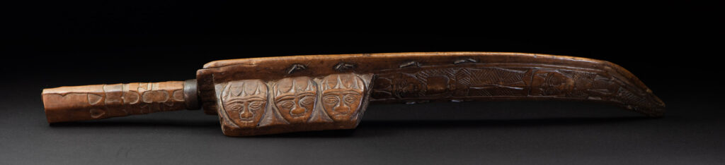 Paiwan tribe hunting knife with a sheath carved with a standing figure and human faces on the side. The hilt is made of carved wood.