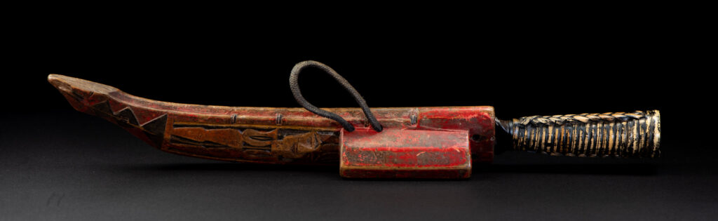 Paiwan tribe hunting knife with a sheath carved with a standing figure and painted in red. The blade and handle are forged as one piece, with the grip wrapped in rattan for protection.