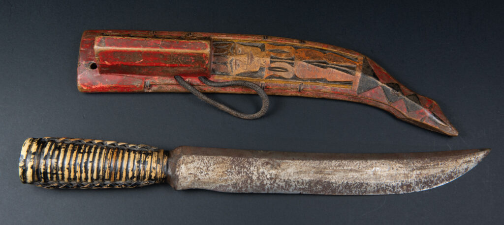Paiwan tribe hunting knife with a sheath carved with a standing figure and painted in red. The blade and handle are forged as one piece, with the grip wrapped in rattan for protection.