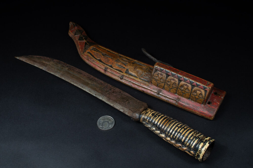 Paiwan tribe hunting knife with a sheath carved with a standing figure and painted in red. The blade and handle are forged as one piece, with the grip wrapped in rattan for protection.