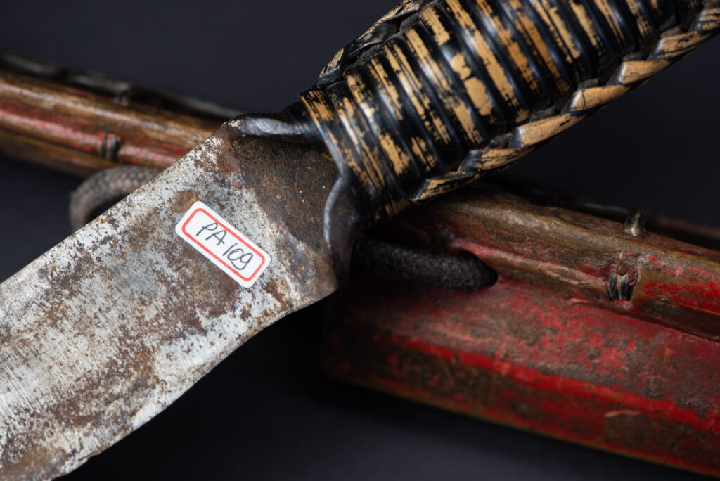 Paiwan tribe hunting knife with a sheath carved with a standing figure and painted in red. The blade and handle are forged as one piece, with the grip wrapped in rattan for protection.
