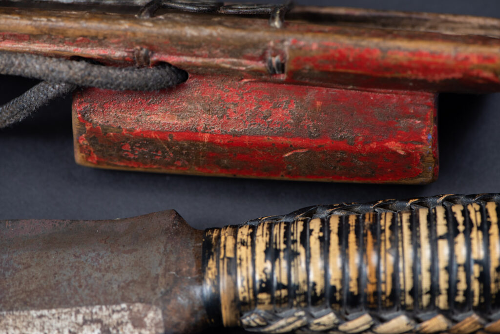 Paiwan tribe hunting knife with a sheath carved with a standing figure and painted in red. The blade and handle are forged as one piece, with the grip wrapped in rattan for protection.