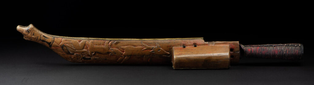 Hunting sword from the Paiwan tribe, with a sheath carved with a scene of a hunter accompanied by his dog shooting at deer. The sides and top of the sheath are covered with brass sheets finely embossed with human head motifs. The sword is made in one piece with the handle, which is wrapped in red-stained rattan.