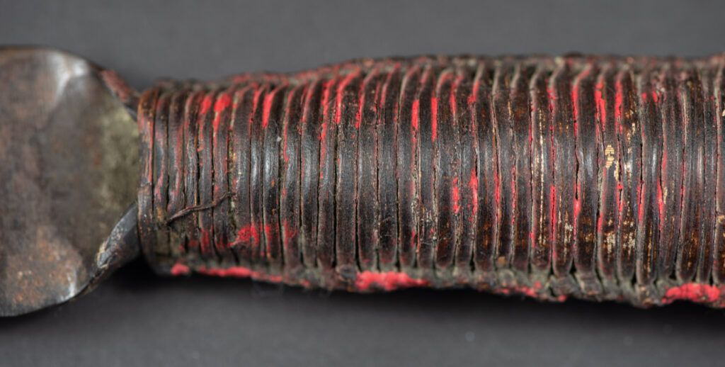 Hunting sword from the Paiwan tribe, with a sheath carved with a scene of a hunter accompanied by his dog shooting at deer. The sides and top of the sheath are covered with brass sheets finely embossed with human head motifs. The sword is made in one piece with the handle, which is wrapped in red-stained rattan.