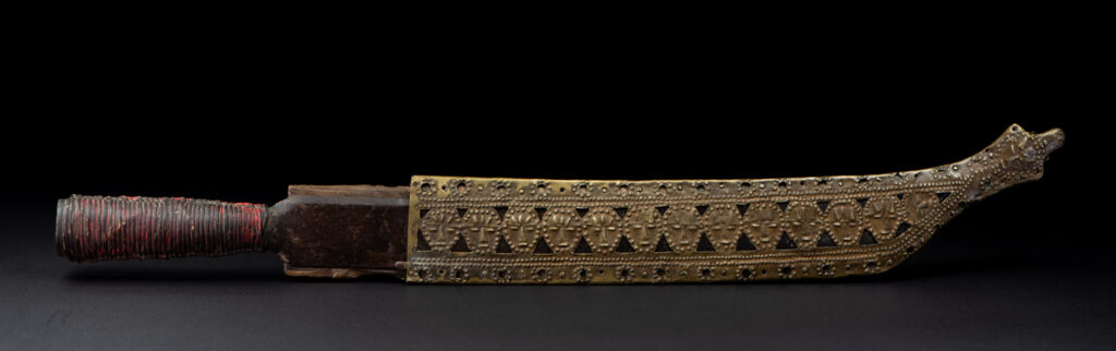 Hunting sword from the Paiwan tribe, with a sheath carved with a scene of a hunter accompanied by his dog shooting at deer. The sides and top of the sheath are covered with brass sheets finely embossed with human head motifs. The sword is made in one piece with the handle, which is wrapped in red-stained rattan.