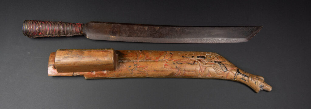 Hunting sword from the Paiwan tribe, with a sheath carved with a scene of a hunter accompanied by his dog shooting at deer. The sides and top of the sheath are covered with brass sheets finely embossed with human head motifs. The sword is made in one piece with the handle, which is wrapped in red-stained rattan.