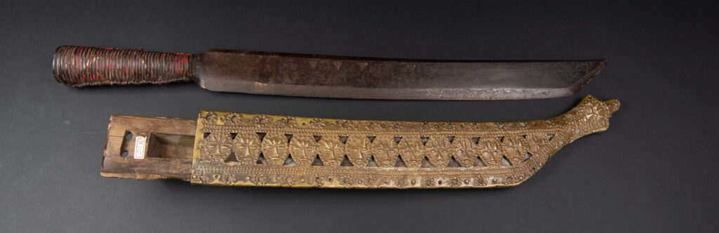 Hunting sword from the Paiwan tribe, with a sheath carved with a scene of a hunter accompanied by his dog shooting at deer. The sides and top of the sheath are covered with brass sheets finely embossed with human head motifs. The sword is made in one piece with the handle, which is wrapped in red-stained rattan.