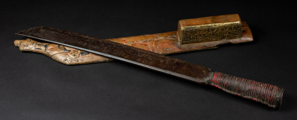 Hunting sword from the Paiwan tribe, with a sheath carved with a scene of a hunter accompanied by his dog shooting at deer. The sides and top of the sheath are covered with brass sheets finely embossed with human head motifs. The sword is made in one piece with the handle, which is wrapped in red-stained rattan.