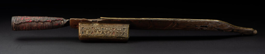 Hunting sword from the Paiwan tribe, with a sheath carved with a scene of a hunter accompanied by his dog shooting at deer. The sides and top of the sheath are covered with brass sheets finely embossed with human head motifs. The sword is made in one piece with the handle, which is wrapped in red-stained rattan.