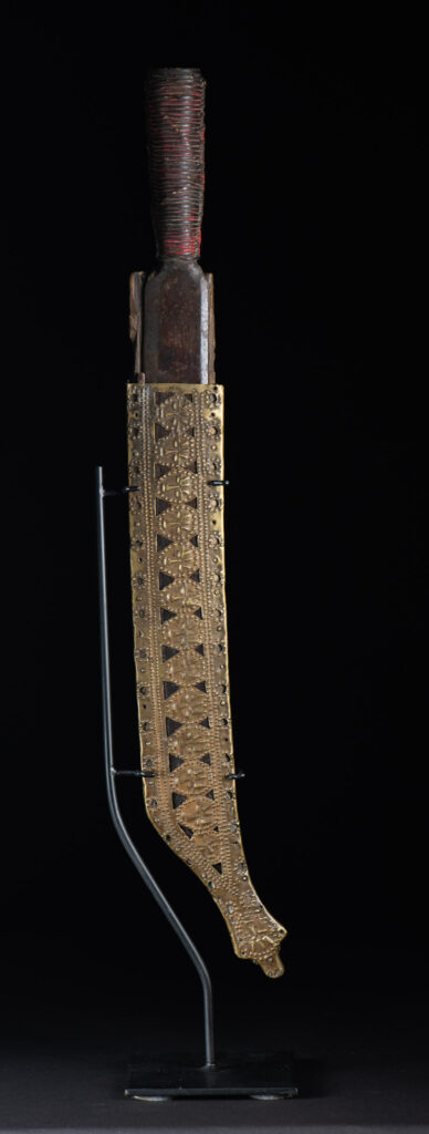 Hunting sword from the Paiwan tribe, with a sheath carved with a scene of a hunter accompanied by his dog shooting at deer. The sides and top of the sheath are covered with brass sheets finely embossed with human head motifs. The sword is made in one piece with the handle, which is wrapped in red-stained rattan.