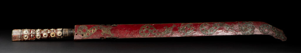Ancient ceremonial long sword from the Rukai tribe. The back of the sheath is painted red and inlaid with finely engraved snakes in brass sheet. The top is also covered with an engraved brass plate. The very long blade of this Rukai sword features a handle in the Rukai style, decorated with small flower-shaped motifs and aluminum wire. The guard of the sword is made from a Japanese coin.