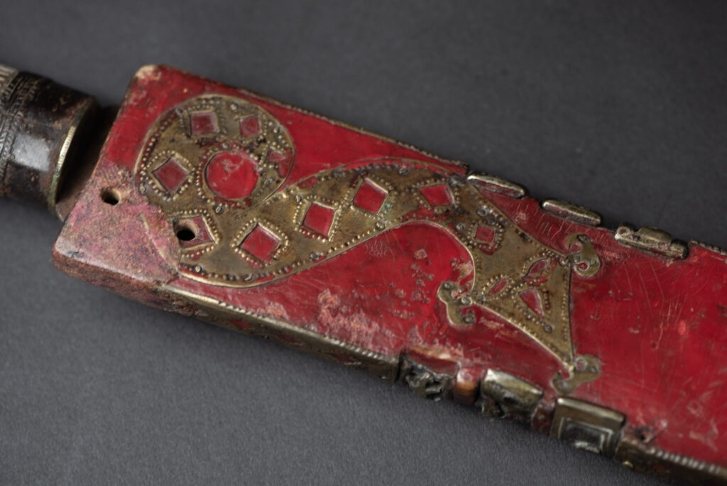 Ancient ceremonial long sword from the Rukai tribe. The back of the sheath is painted red and inlaid with finely engraved snakes in brass sheet. The top is also covered with an engraved brass plate. The very long blade of this Rukai sword features a handle in the Rukai style, decorated with small flower-shaped motifs and aluminum wire. The guard of the sword is made from a Japanese coin.