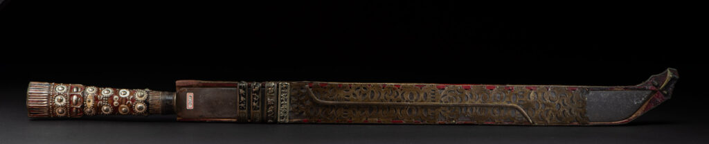 Ancient ceremonial long sword from the Rukai tribe. The back of the sheath is painted red and inlaid with finely engraved snakes in brass sheet. The top is also covered with an engraved brass plate. The very long blade of this Rukai sword features a handle in the Rukai style, decorated with small flower-shaped motifs and aluminum wire. The guard of the sword is made from a Japanese coin.