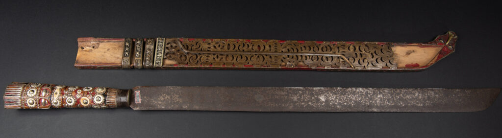 Ancient ceremonial long sword from the Rukai tribe. The back of the sheath is painted red and inlaid with finely engraved snakes in brass sheet. The top is also covered with an engraved brass plate. The very long blade of this Rukai sword features a handle in the Rukai style, decorated with small flower-shaped motifs and aluminum wire. The guard of the sword is made from a Japanese coin.