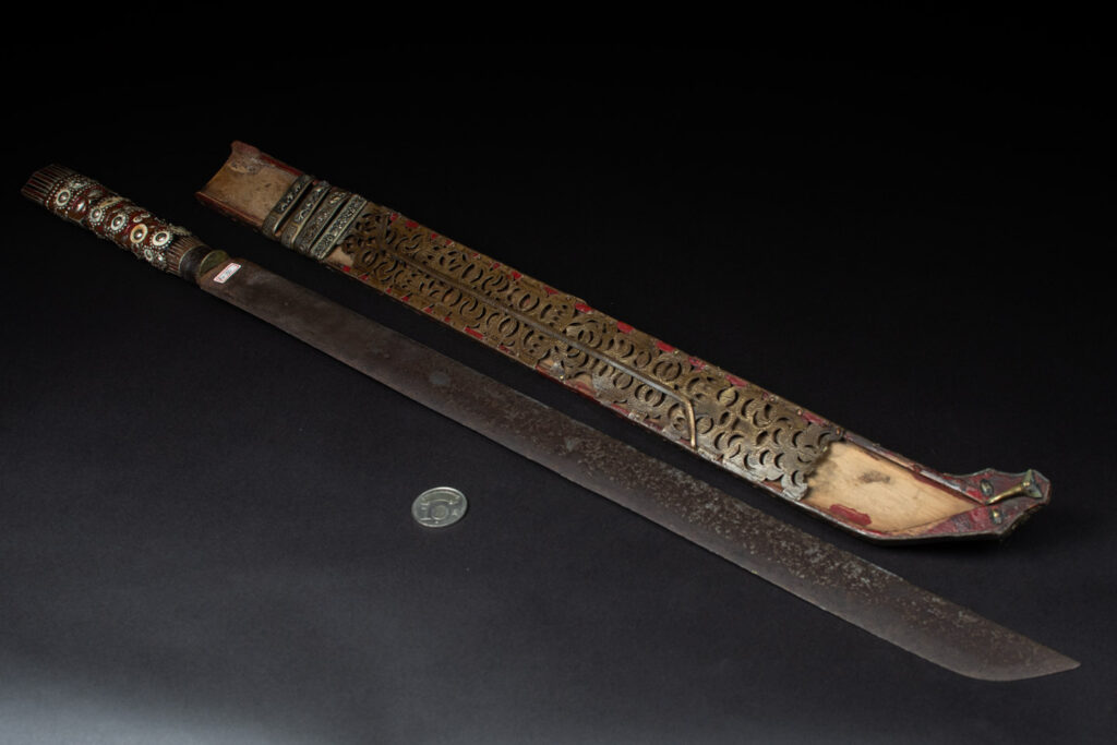 Ancient ceremonial long sword from the Rukai tribe. The back of the sheath is painted red and inlaid with finely engraved snakes in brass sheet. The top is also covered with an engraved brass plate. The very long blade of this Rukai sword features a handle in the Rukai style, decorated with small flower-shaped motifs and aluminum wire. The guard of the sword is made from a Japanese coin.