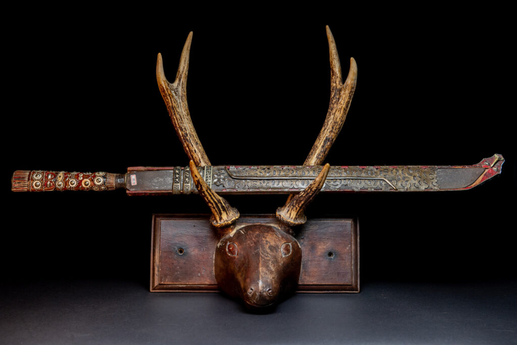 Ancient ceremonial long sword from the Rukai tribe. The back of the sheath is painted red and inlaid with finely engraved snakes in brass sheet. The top is also covered with an engraved brass plate. The very long blade of this Rukai sword features a handle in the Rukai style, decorated with small flower-shaped motifs and aluminum wire. The guard of the sword is made from a Japanese coin.