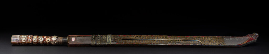 Ancient ceremonial long sword from the Rukai tribe. The back of the sheath is painted red and inlaid with finely engraved snakes in brass sheet. The top is also covered with an engraved brass plate. The very long blade of this Rukai sword features a handle in the Rukai style, decorated with small flower-shaped motifs and aluminum wire. The guard of the sword is made from a Japanese coin.