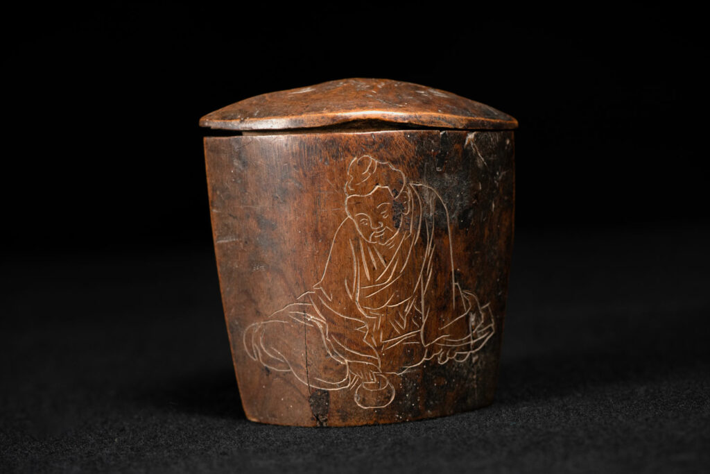 Wooden belt snuffbox from the Pingpu tribes, decorated with an engraving of a Chinese sage. It features a hinged lid carved from a single piece of wood and a section designed for attachment to the belt.