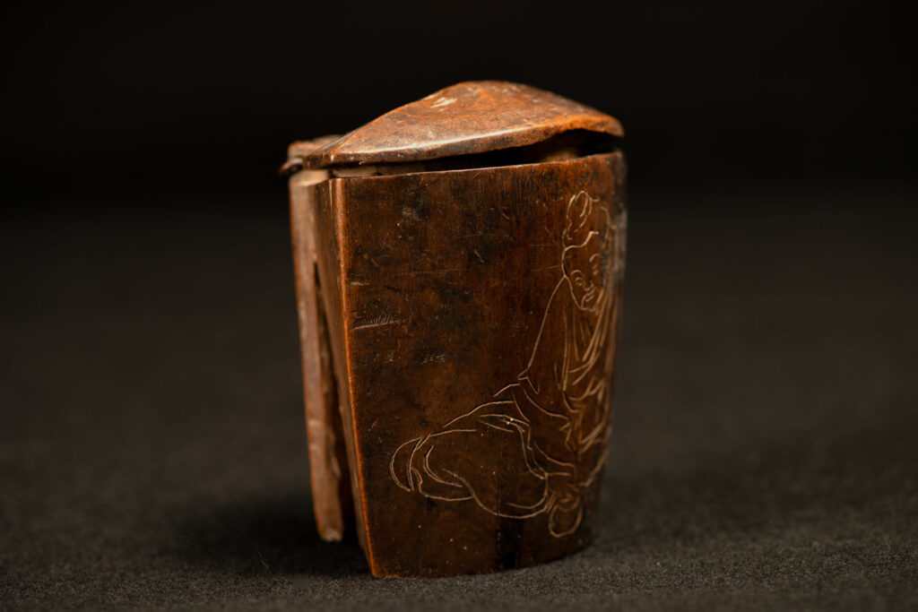 Wooden belt snuffbox from the Pingpu tribes, decorated with an engraving of a Chinese sage. It features a hinged lid carved from a single piece of wood and a section designed for attachment to the belt.